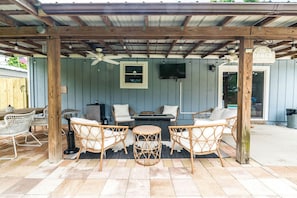 Cozy outdoor lounge with firepit, TV, charcoal grill, dining for 6, mini fridge, fans & string lights — the perfect spot to unwind under the palms