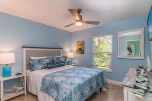 Serene coastal bedroom with queen bed featuring dual nightstands, a ceiling fan, dresser with decor, and a wall-mounted TV.