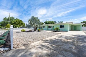 Beach-style home with mint green exterior, shell-themed front door, white picket fence, rock landscaping, palm trees, and a wide driveway.