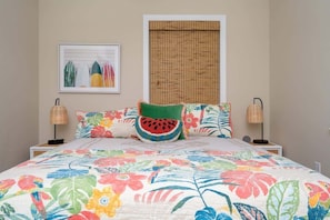 Bright and cheerful bedroom with a king bed, ceiling fan, wall-mounted TV, and double closet. Colorful bedding, fun accent pillows, and woven blinds add a playful tropical touch.