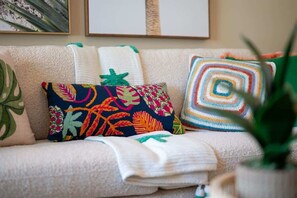 Cheerful living space with a sofa, accent chairs, wicker coffee table, and bar seating. Features coastal artwork, colorful pillows, a smart TV, and a ceiling fan—all giving a beach-inspired setting.
