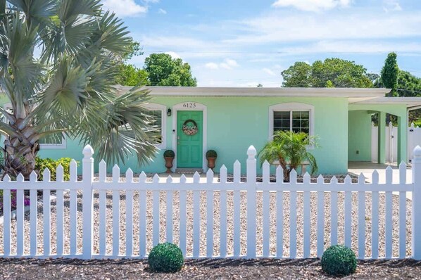 Beach-style home with mint green exterior, shell-themed front door, white picket fence, rock landscaping, palm trees, and a wide driveway.