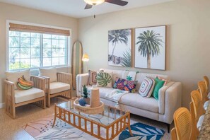 Cheerful living space with a sofa, accent chairs, wicker coffee table, and bar seating. Features coastal artwork, colorful pillows, a smart TV, and a ceiling fan—all giving a beach-inspired setting.
