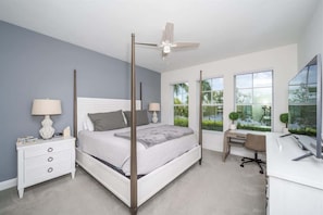 Spacious bedroom with four-poster king bed, matching nightstands, and a large dresser with TV. Featuring large windows and a workspace.