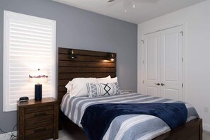 Modern bedroom with a queen bed, wood-paneled headboard, and reading lights. Features a comforter, nightstands, TV, ceiling fan, and direct en-suite access.