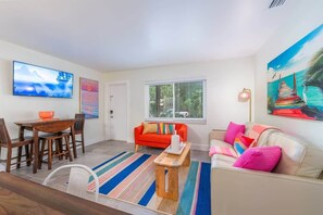 Living area with a leather sofa, bright accent chairs, and a wall-mounted TV. Includes a dining nook, workspace, and open kitchen.