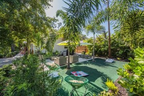 Backyard with wicker lounge seating, lime umbrellas, and lush greenery. Includes shaded loungers, a cozy firepit setup, and cornhole lawn games.