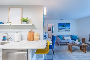 Open-plan living space with a full kitchen, breakfast bar, and dedicated workspace. Features modern appliances, comfy seating, and beachy decor throughout. Includes a mounted TV, dining nook, and vibrant accent furniture.