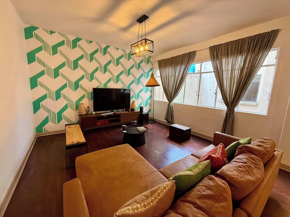 Sala (Vista desde el sofa, un sofa, un cofre, un banco largo, una lampara junto a la tele, televisión con mueble, centro de mesa, alexa, focos, cortinero)