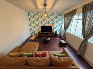 Sala (Vista desde el sofa, un sofa, un cofre, un banco largo, una lampara junto a la tele, televisión con mueble, centro de mesa, alexa, focos, cortinero)