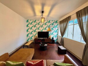 Sala (Vista desde el sofa, un sofa, un cofre, un banco largo, una lampara junto a la tele, televisión con mueble, centro de mesa, alexa, focos, cortinero)