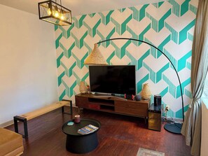 Sala de entretenimiento (visto desde el sofá, un cofre, un banco largo, una lampara junto a la tele, televisión con mueble, centro de mesa, alexa, focos, cortinero)