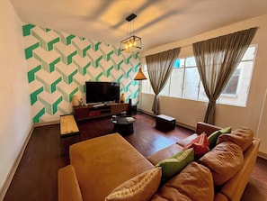 Sala (Vista desde el sofa, un sofa, un cofre, un banco largo, una lampara junto a la tele, televisión con mueble, centro de mesa, alexa, focos, cortinero)