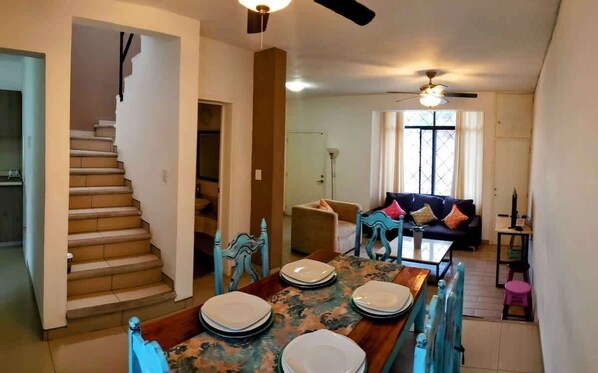 Comedor (visto desde la esquina, mesa con platos y silla, sillones, entrada a la cocina, escalera a los cuartos, ventanas)