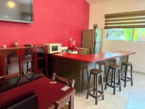 Televisión, vitrina de madera, decoraciones, controles de tv y aire acondicionado, barra, bancos de madera, mesa de madera con vidrio, sillas, microondas, estufa, licuadora, tarja, refrigerador, persianascafés