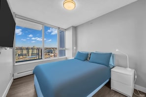 Tranquil and inviting, this bedroom features a plush bed dressed in calming blue tones, large windows offering breathtaking city views, and sleek, minimalist furnishings for a peaceful retreat.