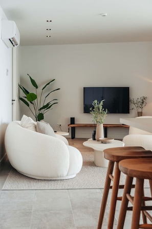 A contemporary living room with soft, sculptural seating, a minimalist coffee table, and a sleek TV setup. Natural light floods the space, creating a peaceful, inviting ambiance