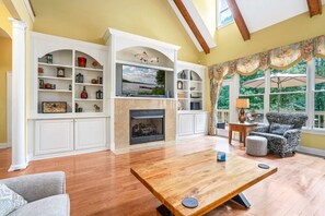Sink into the cozy sofa and relax as sunlight pours through the tall windows, the fireplace flickers, and wooded views create a peaceful backdrop—perfect for unwinding in comfort.
