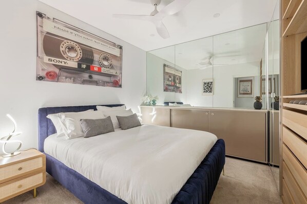 Rest easy in the queen-size bed with soft linens and plush pillows, framed by mirrored storage and artistic touches that add personality and warmth to the bedroom.