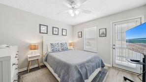 This serene bedroom features a cozy king-size bed, calming coastal décor, and a wall-mounted TV for winding down after a day in the sun. Natural light pours in through the windows and glass door, which leads directly to the backyard. Thoughtful touches li