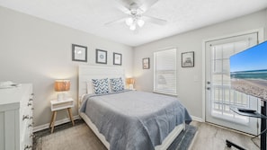 This serene bedroom features a cozy king-size bed, calming coastal décor, and a wall-mounted TV for winding down after a day in the sun. Natural light pours in through the windows and glass door, which leads directly to the backyard. Thoughtful touches li