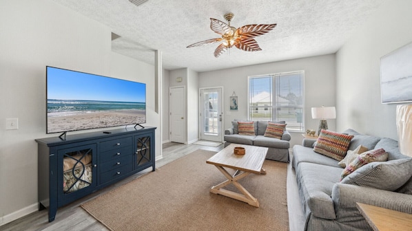 This bright and inviting living room offers a relaxing coastal vibe with plenty of natural light and beachy décor. Cozy seating and a large flat-screen TV make it the perfect spot to unwind after a day at the beach. The tropical ceiling fan and light-tone