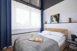 A cozy bedroom with a gray bedspread, sheer curtains, and a painting above the bed. A small nightstand sits next to the bed.