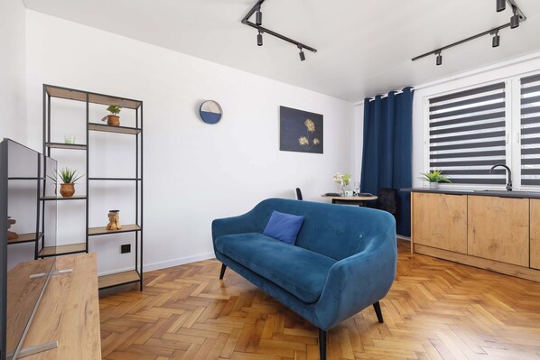 A bright living room with a turquoise armchair, a TV mounted on a stand, and a shelf unit with books and decor. Natural light fills the space.