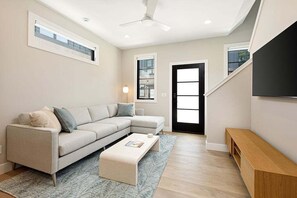Relax in the stylish living room featuring a spacious sectional sofa and a large smart TV—perfect for movie nights or catching up on your favorite shows. The modern design and natural light create a welcoming atmosphere.