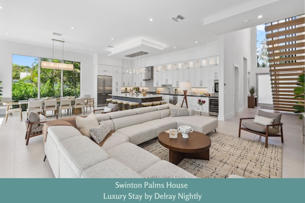 Bright & Modern Delray Beach Retreat – Spacious open-concept living with a designer kitchen, large island, and floor-to-ceiling windows showcasing lush tropical views.