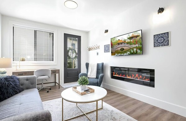 Living room with big screen smart tv, electric fireplace, office desk, window.