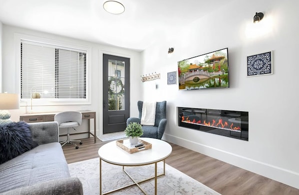 Living room with big screen smart tv, electric fireplace, office desk, window.