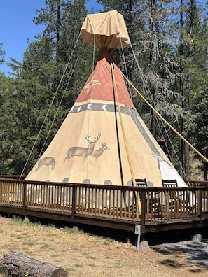 26-foot authentic tipi on an oversized deck with cedar railing—outdoor rocking chairs. Towering lodgepoles rise 30 feet overhead, draped in hand-painted canvas that glows amber at sunrise and flickers gold by the firelight at night.