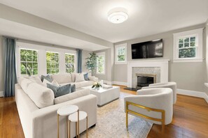 Relax in the airy living room featuring a smart 65-inch Roku TV, two plush sofas and modern chairs that fit 10, hardwood floors, and plenty of natural light from large windows.