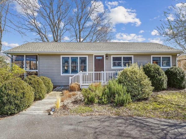 This charming single-story home features a welcoming front porch surrounded by lush greenery and a warm, inviting entryway with large windows offering a view of the sky.