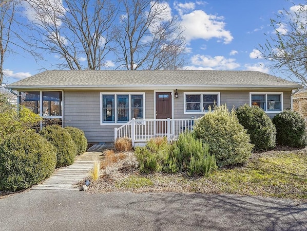 This charming single-story home features a welcoming front porch surrounded by lush greenery and a warm, inviting entryway with large windows offering a view of the sky.