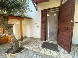 Ingresso rustico ombreggiato da un ulivo, con zanzariera e pavimento in pietra.