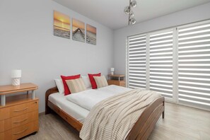A cozy bedroom with red-accented bedding, light wood furniture, and large horizontal blinds allowing natural light in.