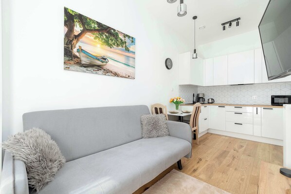 A cozy living room corner with a light gray sofa, wall-mounted TV, and a painting above the couch next to an open-plan kitchen.