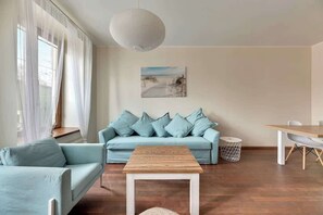 The room’s spaciousness is accentuated by tall windows framed with sheer curtains. A modern TV and minimalist decor enhance the contemporary aesthetic.