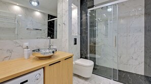 A modern bathroom featuring a glass walk-in shower, marble-style tiles, and a wooden vanity unit with a built-in washing machine for convenience.