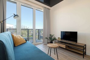 A living area with a sofa and yellow accent pillow, positioned in front of a TV and balcony access.