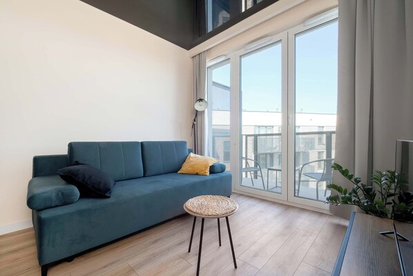 A bright living room with a teal sofa, round wooden coffee table, and floor-to-ceiling window leading to a balcony.