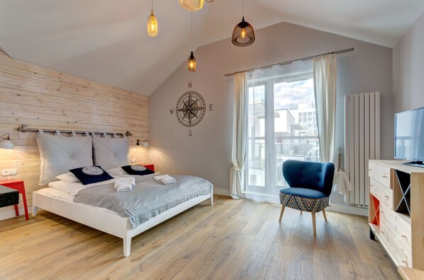 A cozy studio with a nautical theme, featuring a bed, sitting area, and large windows letting in natural light.
