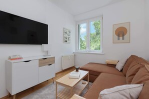 A living room furnished with a brown corner sofa, white TV stand, and large windows letting in natural light.
