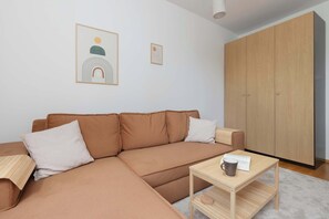 A snug living room with a chaise-style sofa and light wood furnishings, adjacent to a wardrobe.

