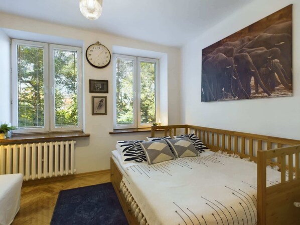A cozy bedroom with a large double bed, wooden floors, and bright natural light coming from two windows. The decor is simple, with a blue rug and an elegant painting on the wall.