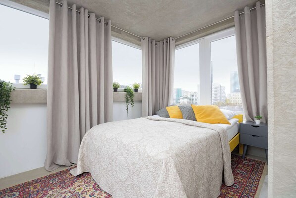 A bright bedroom with a double bed, large windows, beige curtains, and a cozy, minimalistic decor.