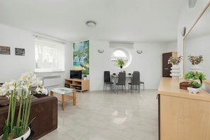 An open-plan living and dining area featuring a brown sofa, potted plants, and a light wooden cabinet.