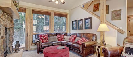 A cozy living room with a brown leather sectional sofa, red ottoman, stone fireplace, and wooden accents. Large windows provide ample natural light, and a staircase is visible in the background.